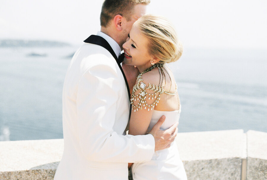 Photographer in Italy: An exquisite and luxurious couple of newlyweds who are hugging on the background of the coast in Monaco. Groom in a white tuxedo, takes his smiling bride, who is dressed in a designer wedding dress decorated with stones