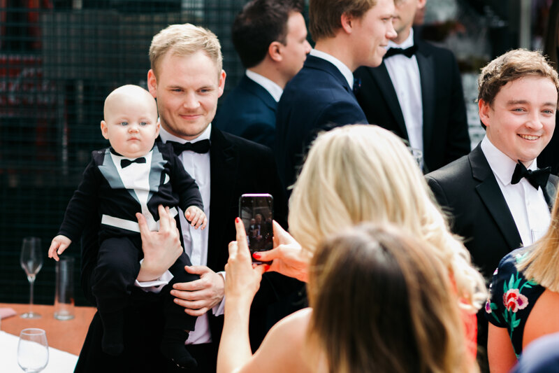 Wedding photography portfolio in Italy: A man holds a little boy in a black tuxedo in his arms, and a woman photographs them on the phone