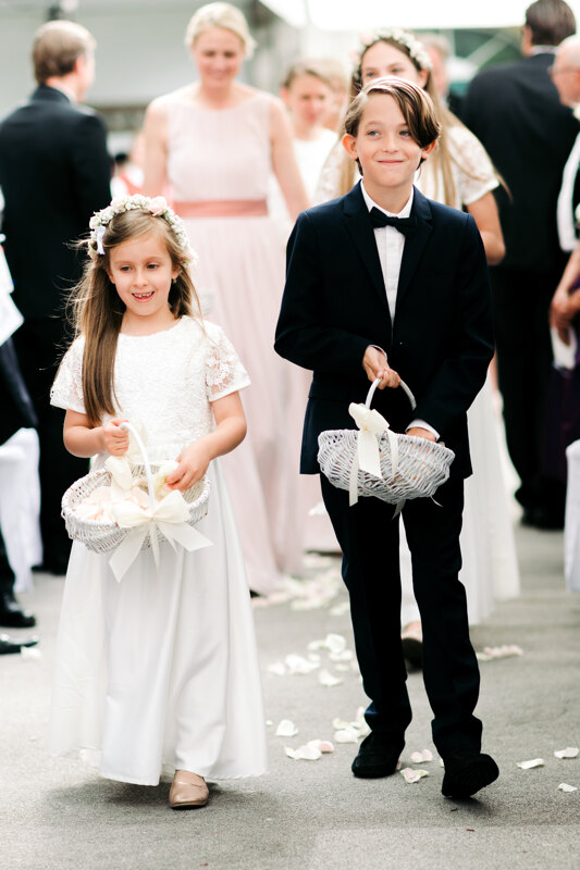 Wedding photography portfolio in Italy: A young man in a tuxedo and a girl in a white dress with a wreath on his head throw rose petals along the path from their white baskets