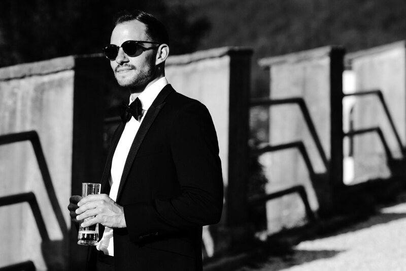 Wedding photography portfolio in Italy: Handsome man in a black suit and glasses holding a glass in his hands