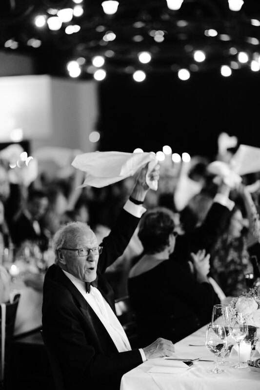 Wedding photography portfolio in Italy: Grandfather in a black tuxedo sits at a table and gaily waves a white napkin over his head