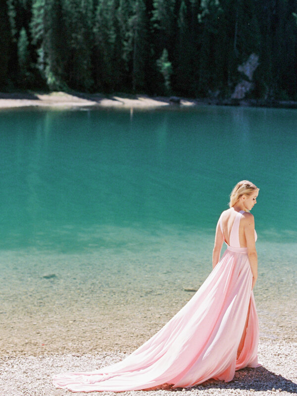 Wedding photography portfolio in Italy: An incredibly beautiful bride in a delicately pink dress stands on the shore of a mountain lake in the Dolomites