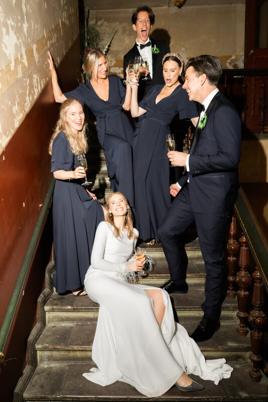 Wedding photography portfolio in Italy: Newlyweds with friends posing for a photographer and drinking champagne on the restaurant stairs