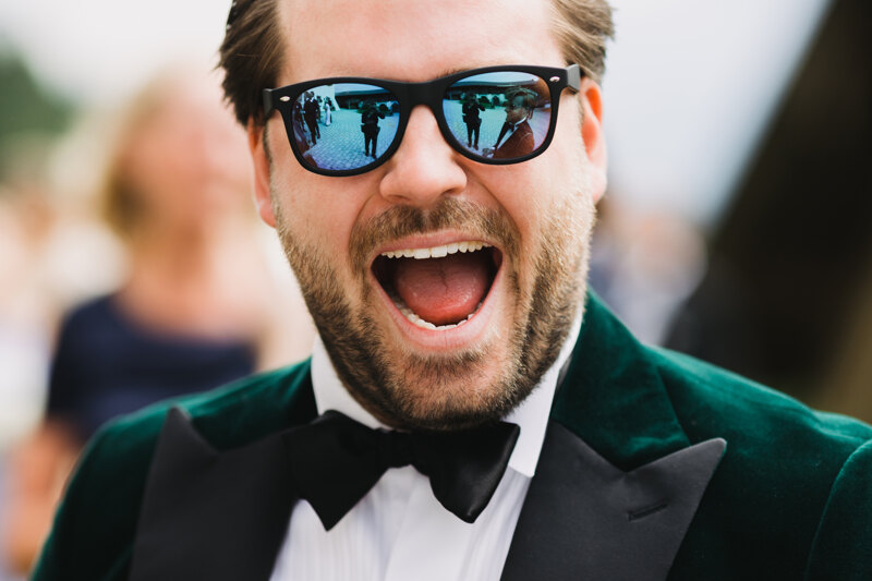 Wedding photoshoot in Italy: Portrait of a groom in colored glasses, dressed in a green tuxedo with a bow tie on his neck and smiles broadly