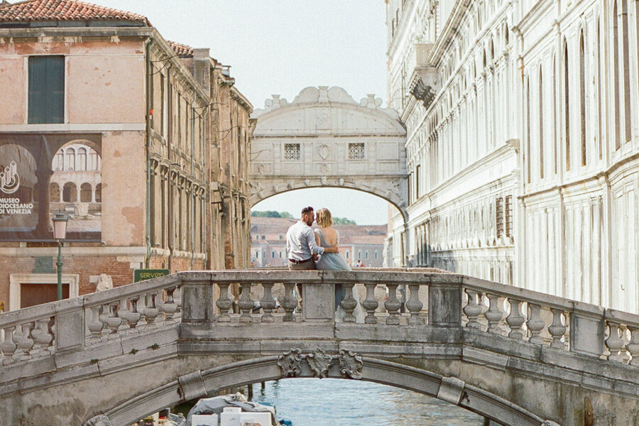 Services of Wedding Photographer in Italy: A beautiful, young couple sits and gently hugs on one of the bridges of romantic and sunny Venice