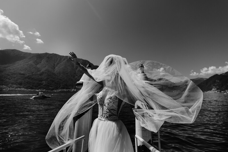 Wedding photography portfolio in Italy: A bride in a white wedding dress, trying to curb her wedding veil, which is strongly dispelled by the wind, on the lake, among the mountains