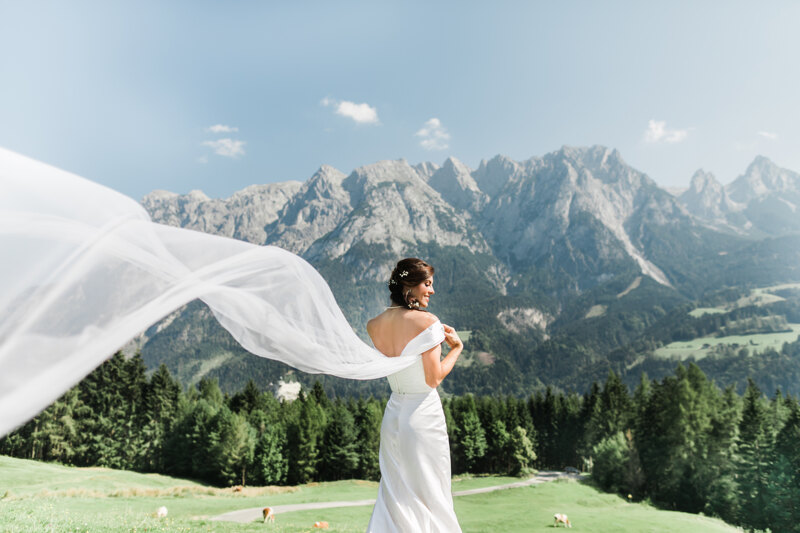 Wedding photography portfolio in Italy: A fragile and slender bride stands on a background of green forest and high mountains, in a white dress with a long train flying in the wind