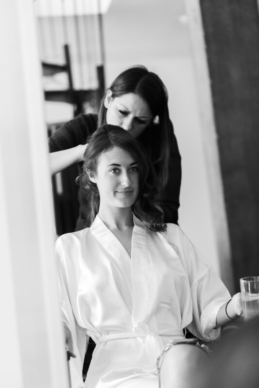 Wedding photography portfolio in Italy: A bride in a white dressing gown with a glass of water in her hand, sitting in a chair in front of a mirror and watching a hairdresser doing her hair