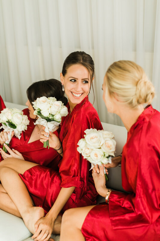 Wedding photography portfolio in Italy: Bridesmaids in red dressing gowns with small bouquets of roses communicate at a morning photo session