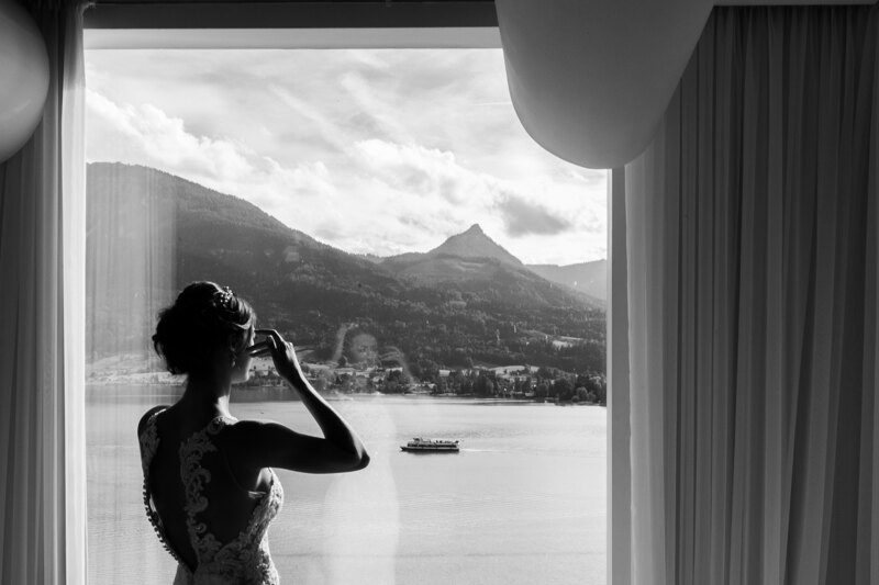 Wedding photography portfolio in Italy: Photo of a dressed bride in her wedding dress, she stands in front of a large window and admires the magnificent view of the mountain lake