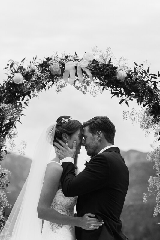 Wedding photography portfolio in Italy: Portrait of the bride and groom, who are standing under the arch of flowers, and the groom gently touches the face of his beloved