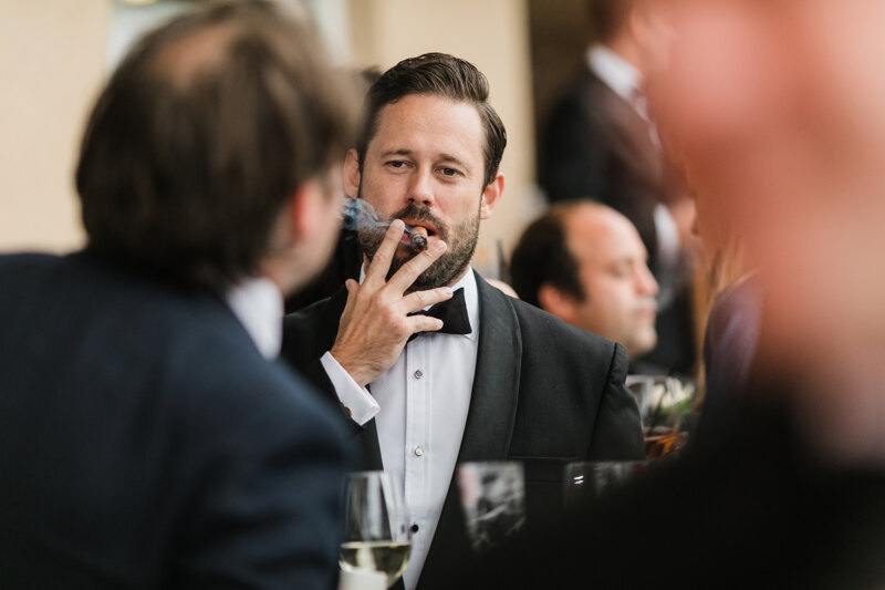 Wedding photography portfolio in Italy: A handsome man in a tuxedo with a neat haircut and beard smokes a cigar